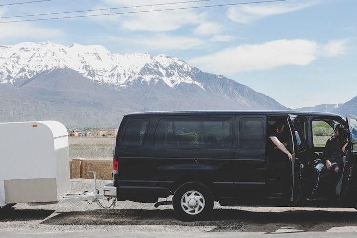Ein schwarzer Tour-Van mit weißem Anhänger vor einer Bergkulisse.